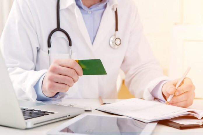 Um médico segura um cartão em uma mão enquanto escreve num papel. A foro está cortada e não é possível ver o rosto dele