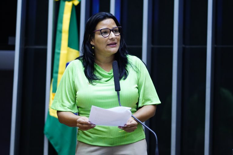 Deputada Juliana Cardoso fala ao microfone. Ela é uma mulher clara, com cabelo escuro, liso e comprido, usa uma blusa verde claro e óculos, e segura um papel