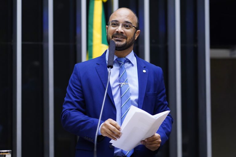 Deputado Capitão Alden fala ao microfone. Ele é um homem claro, usa barba e bigode escuros, é calvo, usa óculos, veste um terno azul e segura um papel